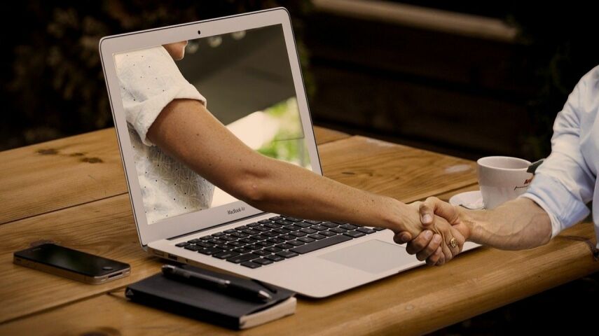 Fotomontaż: ze stojącego na stole laptopa wychodzi mędka ręka ściskająca dłoń mężczyzny stojącego przy biurku