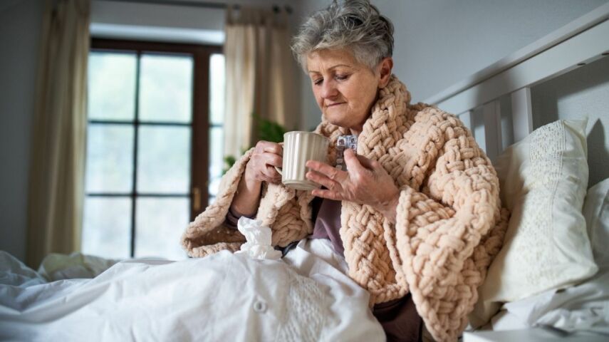W pokoju dla senora z wygodnymi meblami na łóżku siedzi starsza pani okryta ciepłym szlafrokiem pijąc z kubka