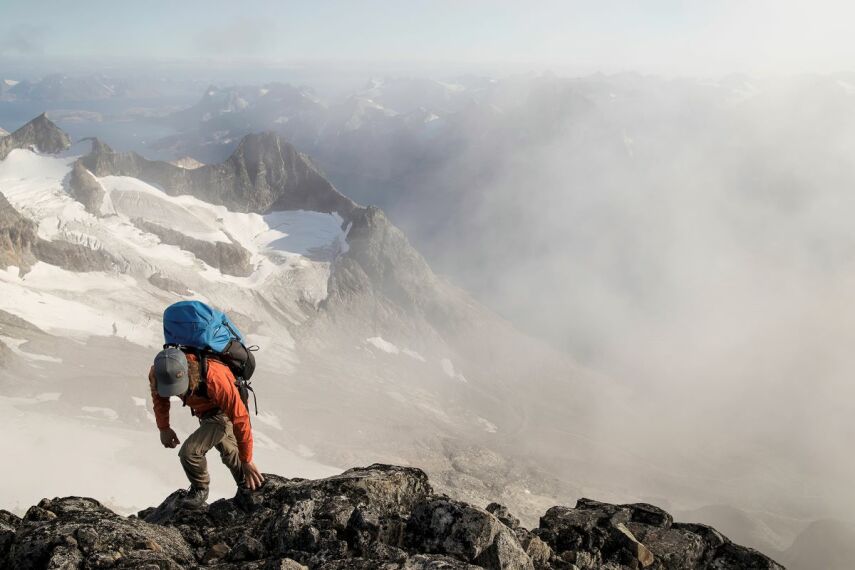 Na skalnej grani z mgłą w tle człowiek z niebieskim plecakiem oraz sprzętem i odzieżą sportową zaawansowaną Arcteryx