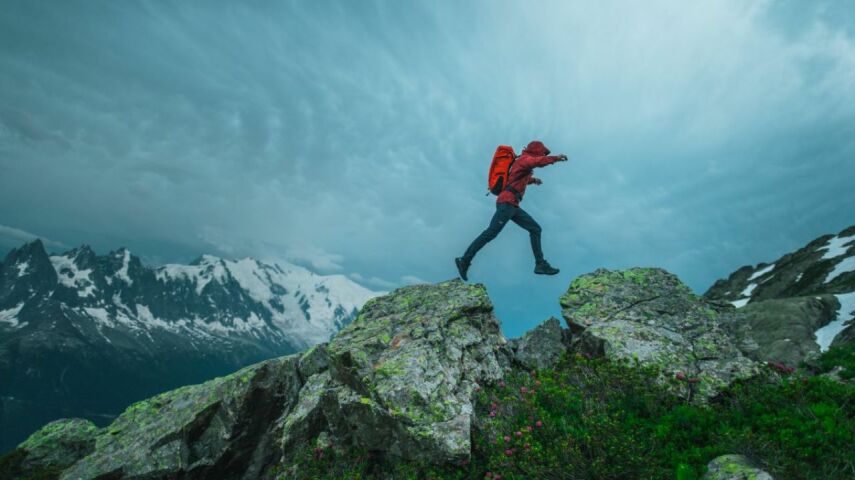 Na szczycie gór mężczyzna w sportowym ubraniu, z plecakiem w obuwiu sportowym Arcteryx przeskakuje z wierzchołka na wierzchołek