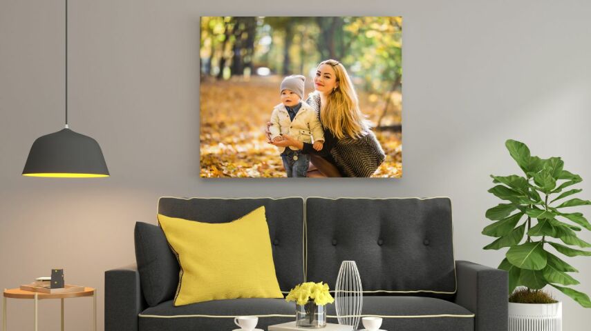 W salonie o szarych ścianach, nadciemnoszarą sofą z żółtymi poduszkami wisi fotobraz z matką tulącą syna w jesiennym krajobrazie