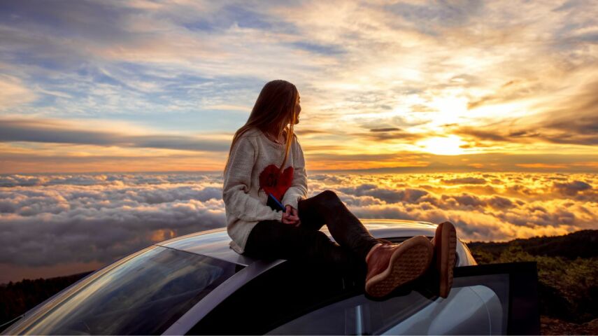 Na dachu samochodu ubezpieczonego online siedzi kobieta wpatrująca się w zachód słońca