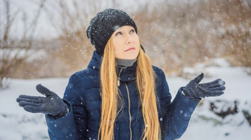 Kobieta w czapce i ciemnej puchowej kurtce zimowej rozkłada ramiona na padający śnieg