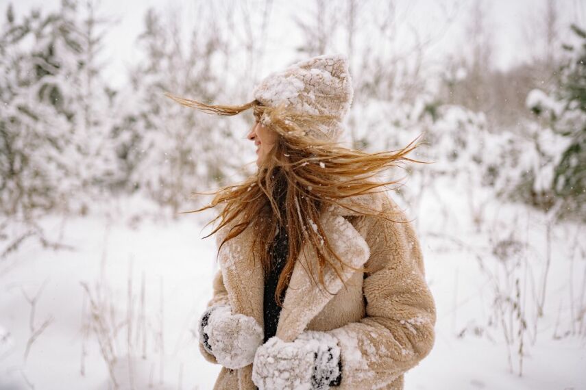 W padającym śniegu radosna kobieta w czapce rękawiczkach i pluszowej beżowej kurtce zimowej