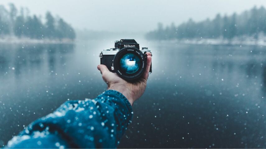 W śniezny dzień na tle jeziora aparat trzymany w ręce skierowany w stronę człowieka